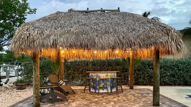 Ein reetgedecktes Gartenhaus mit Tisch und Stühlen, umgeben von einer gepflasterten Terrasse.