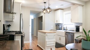 A modern kitchen with white cabinetry, a stainless steel range, a built-in microwave, and a toaster. There is a wooden dining table with chairs, a hanging light fixture, and a potted plant.