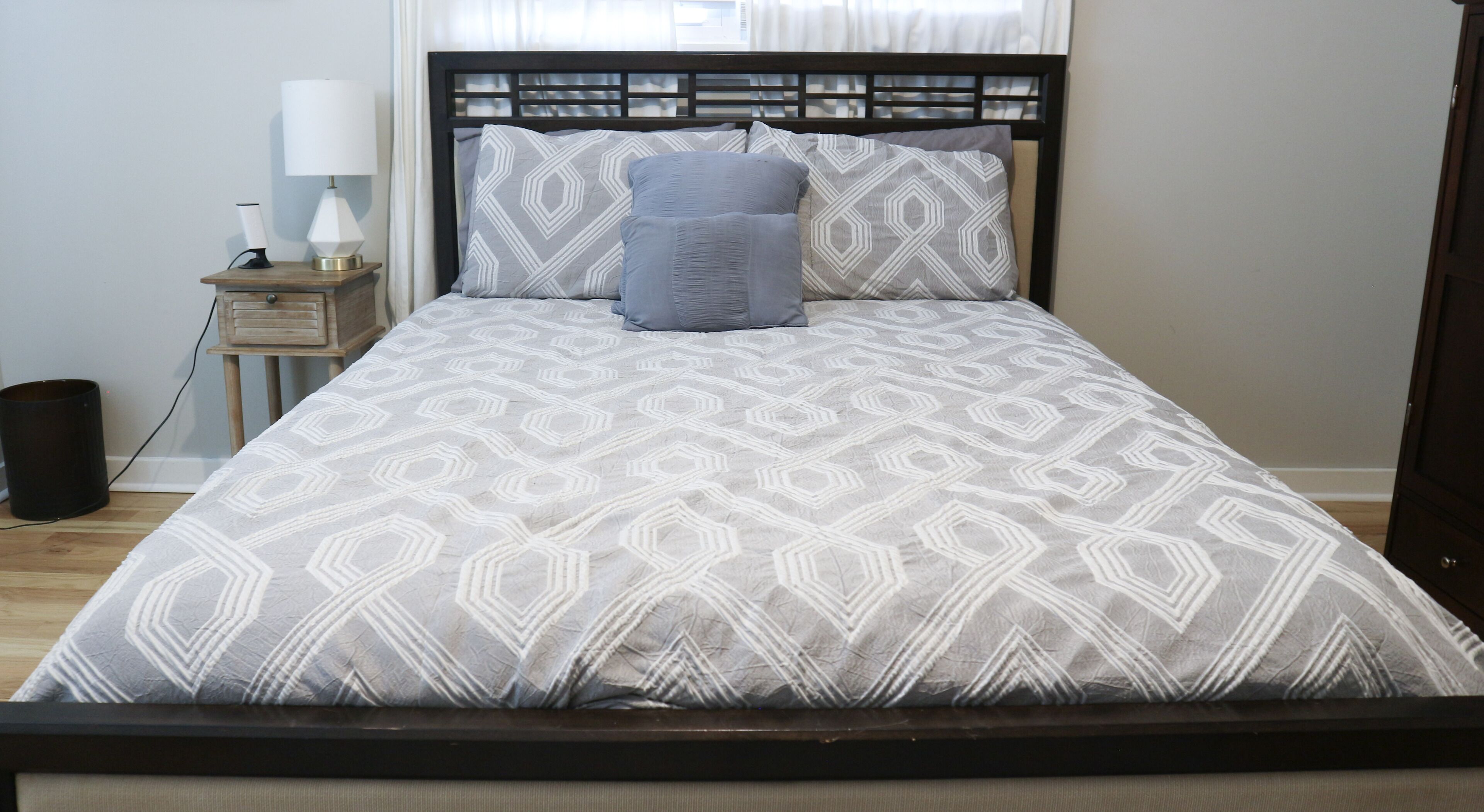 A neatly made bed with a geometric patterned bedspread and matching pillows, a bedside table with a lamp, and a wooden floor.