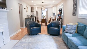 A modern living room with a blue sofa, two leather armchairs, a dining table with chairs, and a kitchen in the background.