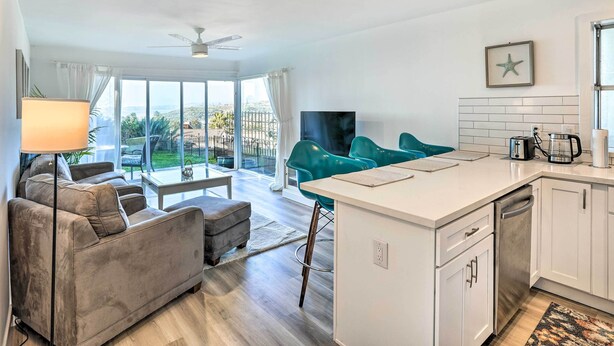 Een moderne woonkamer met een grijze bank, blauwgroene fauteuils, een salontafel en een televisie. Er is een keuken met witte kasten en een klein eiland.