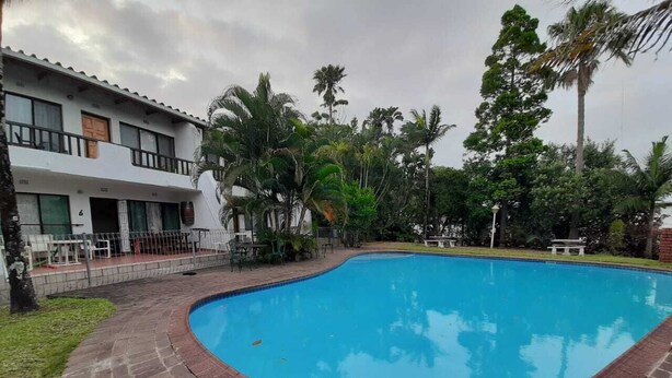 A swimming pool with a brick border, surrounded by palm trees and a building with balconies.
