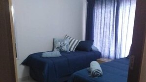 A small, neatly arranged bedroom with two beds, a window with curtains, and a tiled floor.