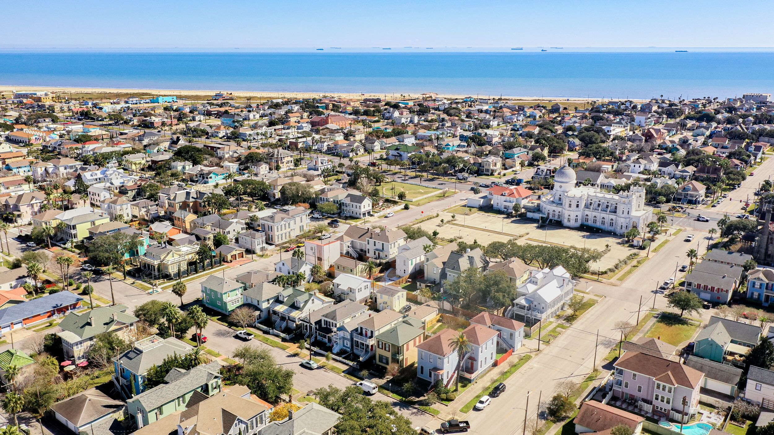 Vista aérea de una zona residencial costera con casas, un gran edificio blanco con cúpula y una playa al fondo.
