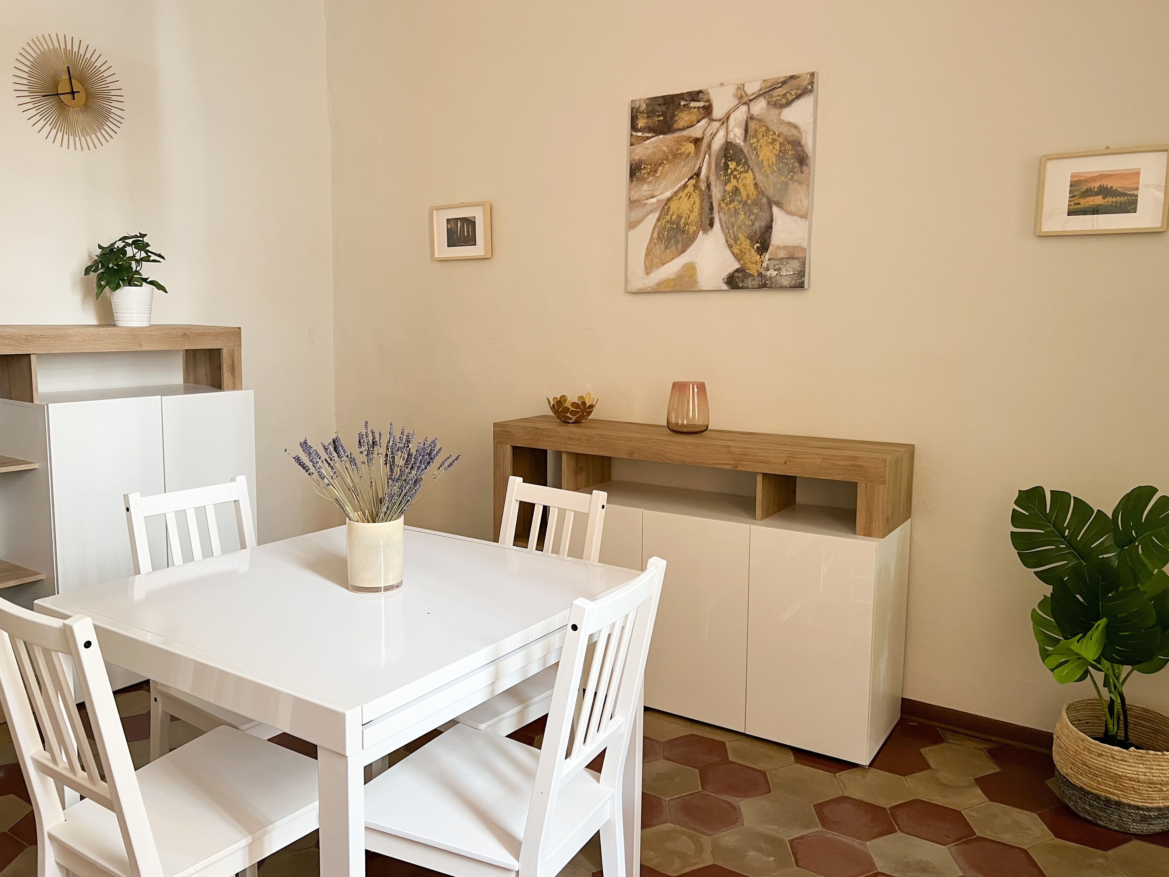 Un comedor con una mesa y sillas blancas, un jarrón de lavanda, un aparador de madera y un cuadro abstracto grande en la pared.