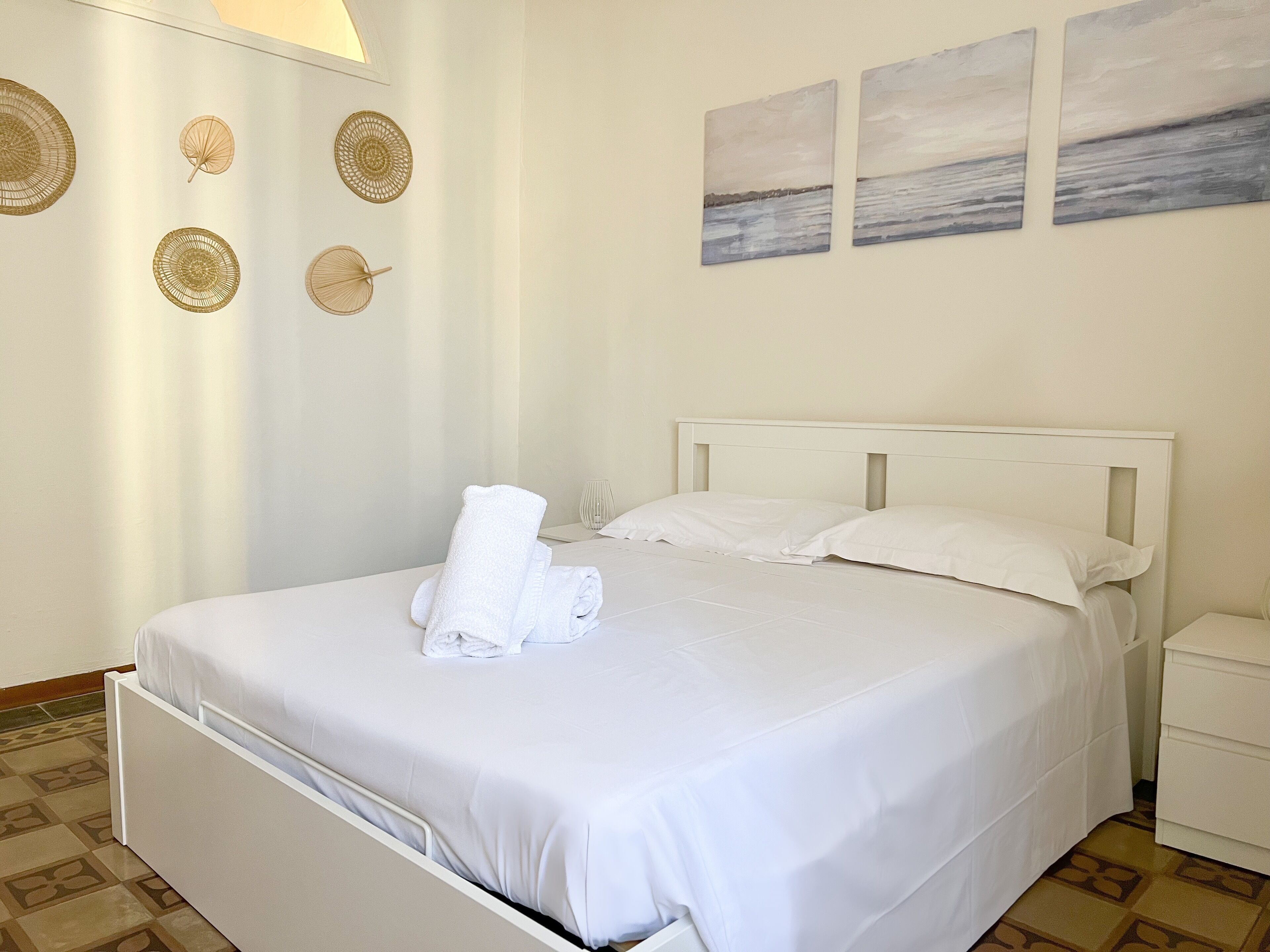 A neatly made bed with white linens and a rolled towel, a bedside table with a glass, and wall decor including circular and rectangular patterns.