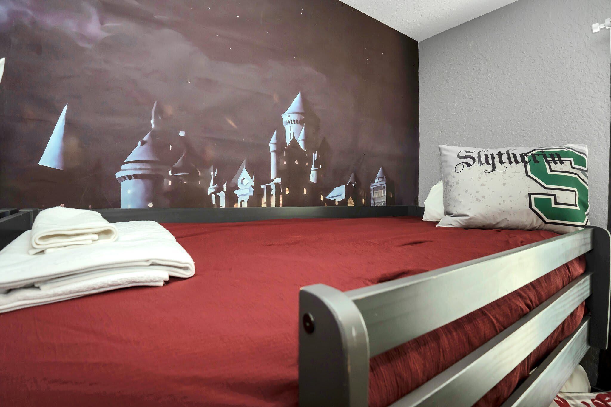 Habitación de hotel con una colcha roja, una toalla blanca doblada con cuidado y una almohada con el texto 'Slytherin'.