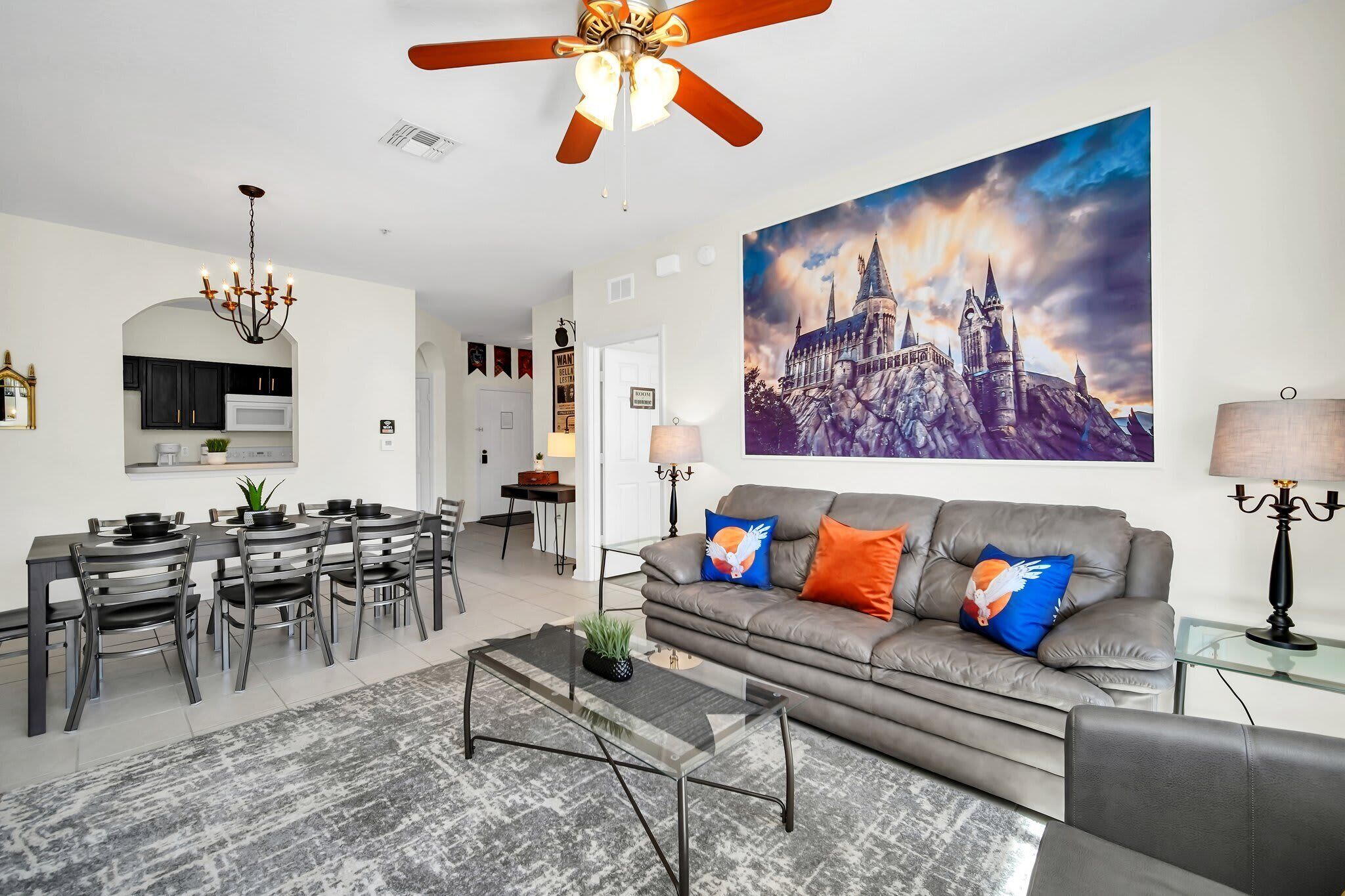 Uma sala de estar moderna com um sofá cinzento, uma mesa de jantar com cadeiras, um candelabro e um quadro grande de um castelo.