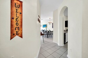 A hallway with a 'WELCOME' sign, a dining area with a table and chairs, and a kitchen with cabinets and a sink.