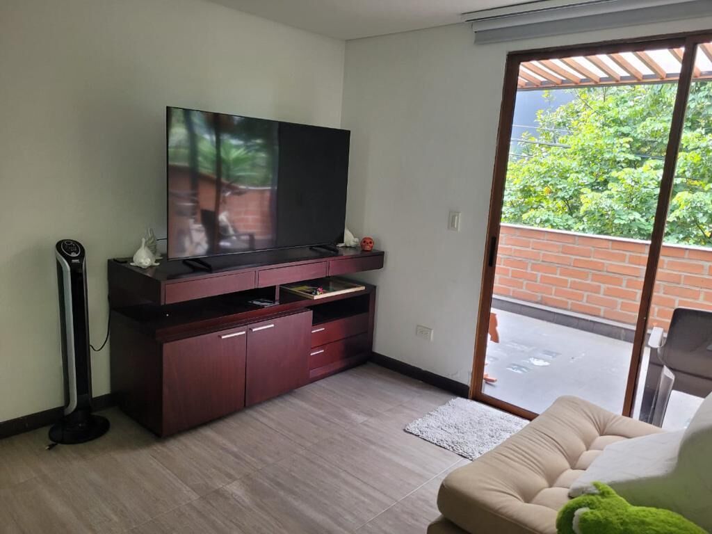 A living room with a flat-screen TV, a dark wooden entertainment unit, a beige sofa, and a sliding glass door leading to a balcony with a brick wall and greenery outside.