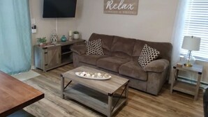 A living room with a brown sofa, a coffee table with decorative items, a TV mounted on the wall, and a window with blinds. - Beach Retreat I HotTub I FirePit I Near Cape May I 7 Beach Passes I 4 Bikes (Lower Township)