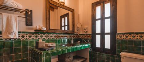 Un baño con pared de azulejos verdes, un estante de madera, un lavabo, un espejo, un perchero para toallas y un inodoro.