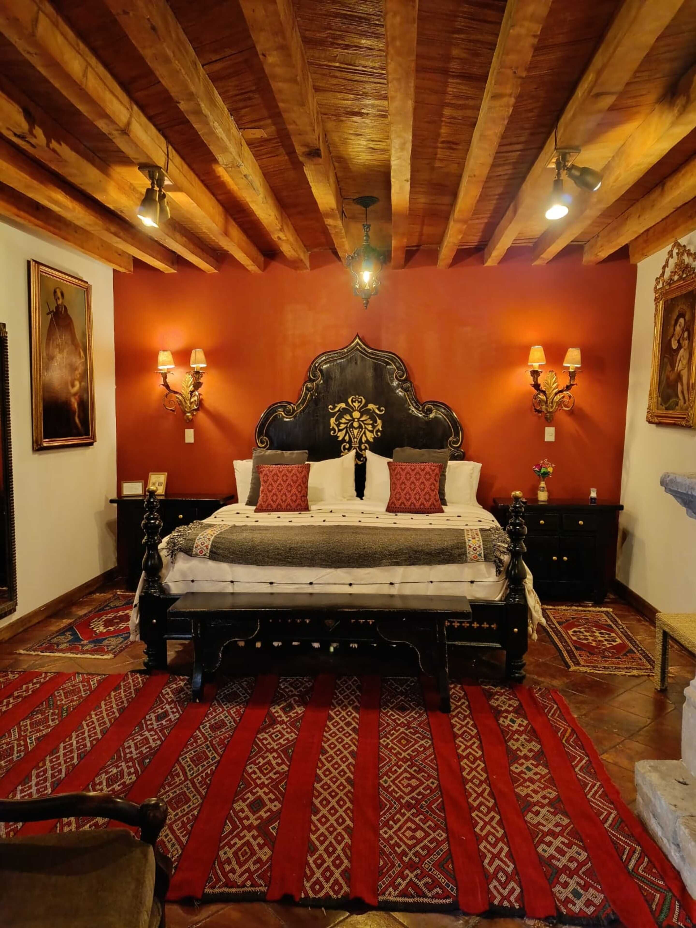 A traditional bedroom with a wooden ceiling, a large bed with ornate headboard, two wall sconces, and patterned rugs.