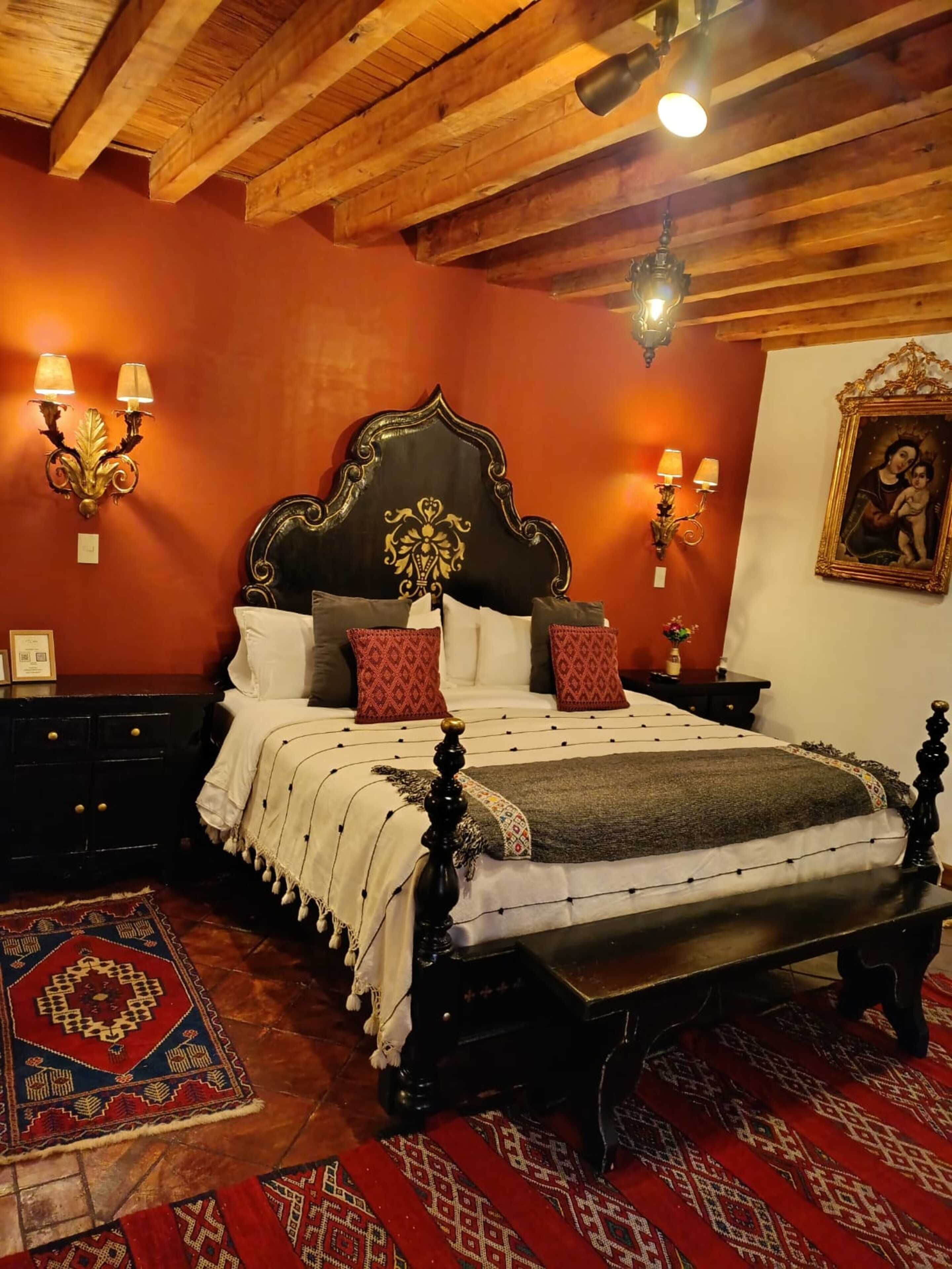 Una habitación tradicional con techo de madera, una cama grande con cabecera oscura, múltiples almohadas y dos lámparas de pared.