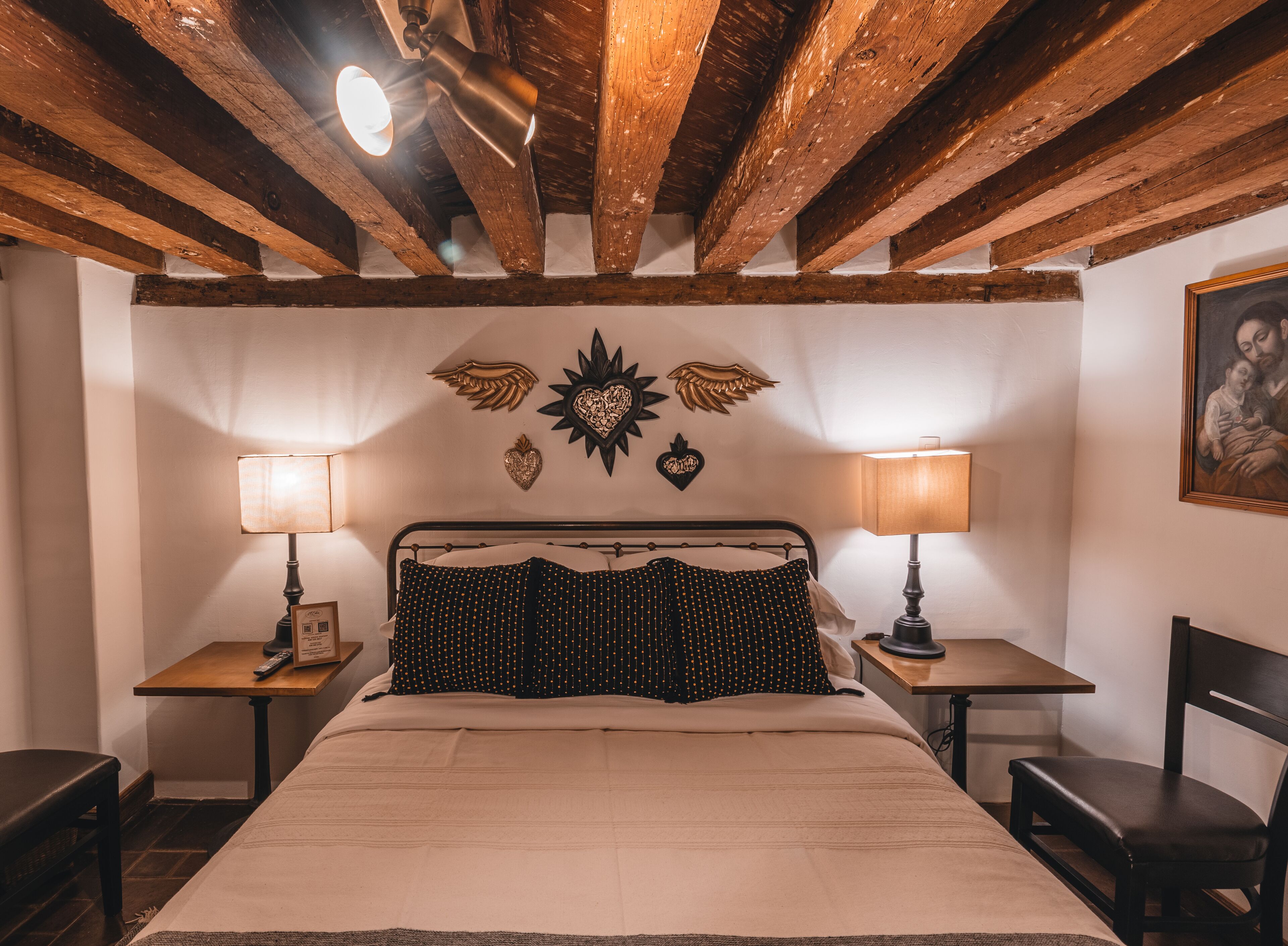 A bedroom with a bed, two bedside tables, a chair, and a ceiling with exposed wooden beams.