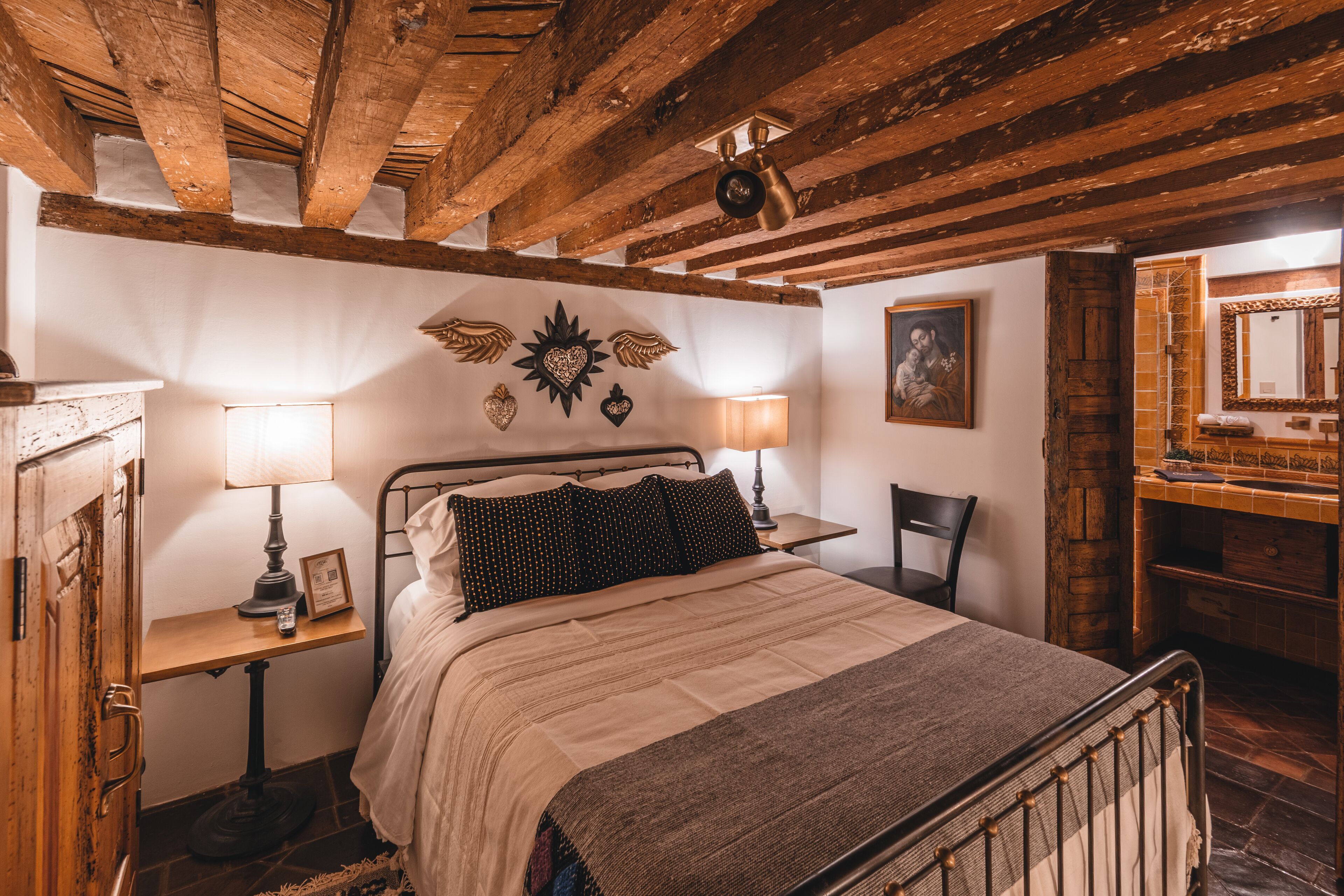 Un dormitorio con techo de madera, una cama con colcha estampada, dos mesitas de noche con lámparas, una silla y un espejo.