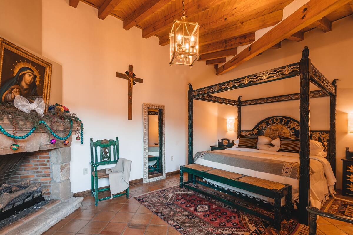 Un lit traditionnel à baldaquin, doté d’une tête de lit en bois, d’une cheminée avec une manteau de cheminée et d’une croix au mur.