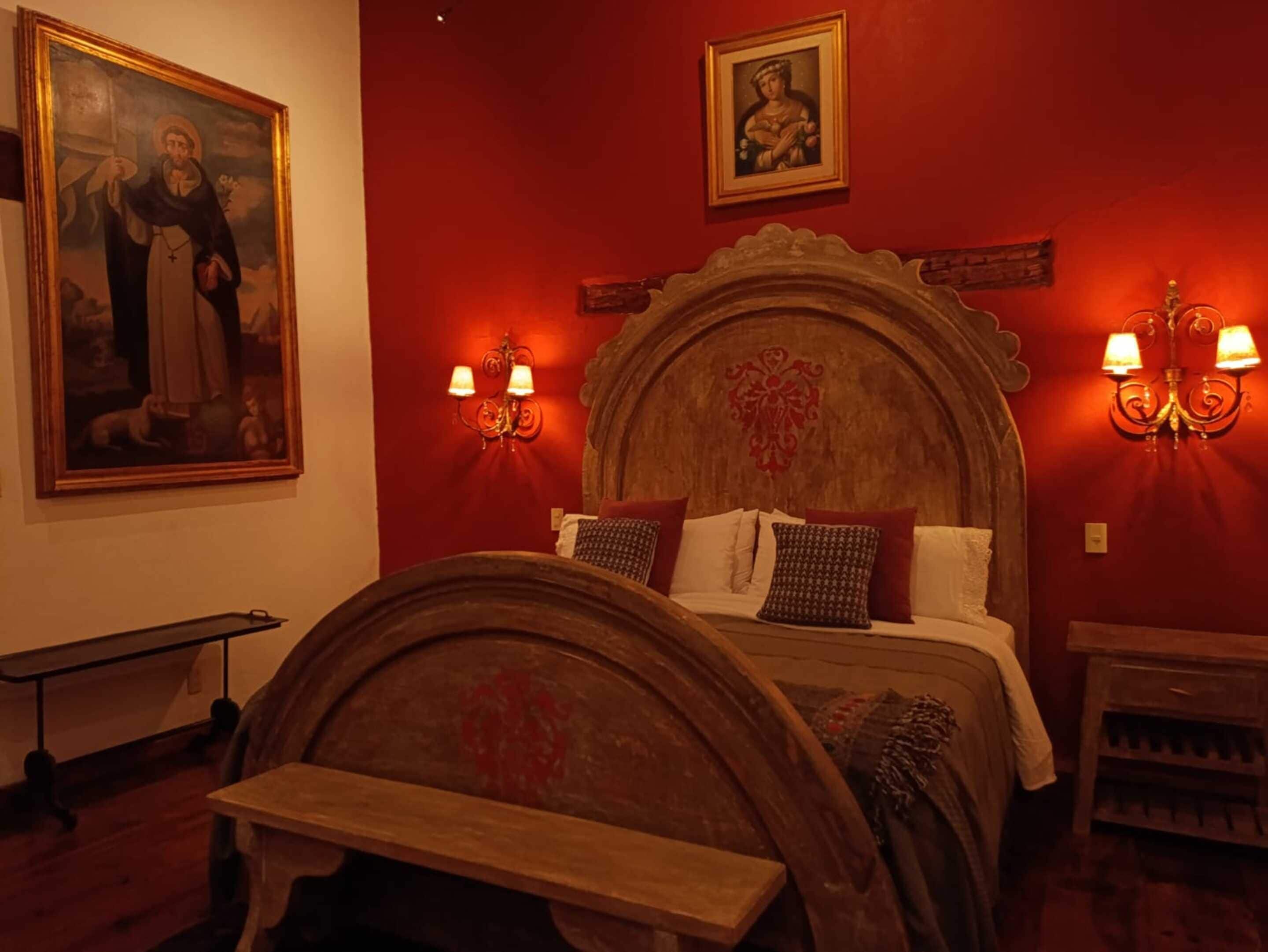 Quarto com cama de madeira, paredes vermelhas, uma pintura grande e duas arandelas.