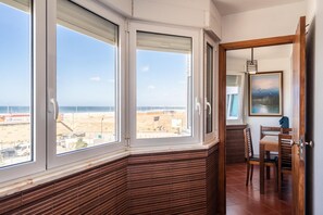 Quarto com grandes janelas com vista para a praia, área de jantar com mobiliário de madeira e um quadro na parede.