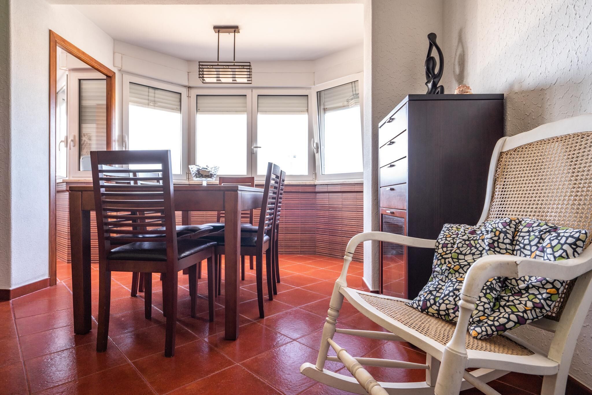 Un comedor con una mesa y sillas de madera, un armario negro y un sillón de mimbre con cojines estampados.