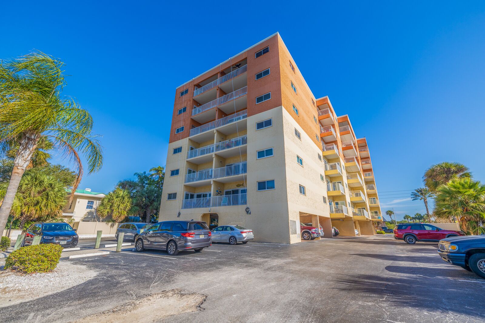 A multi-story hotel with balconies, a parking lot, and palm trees.