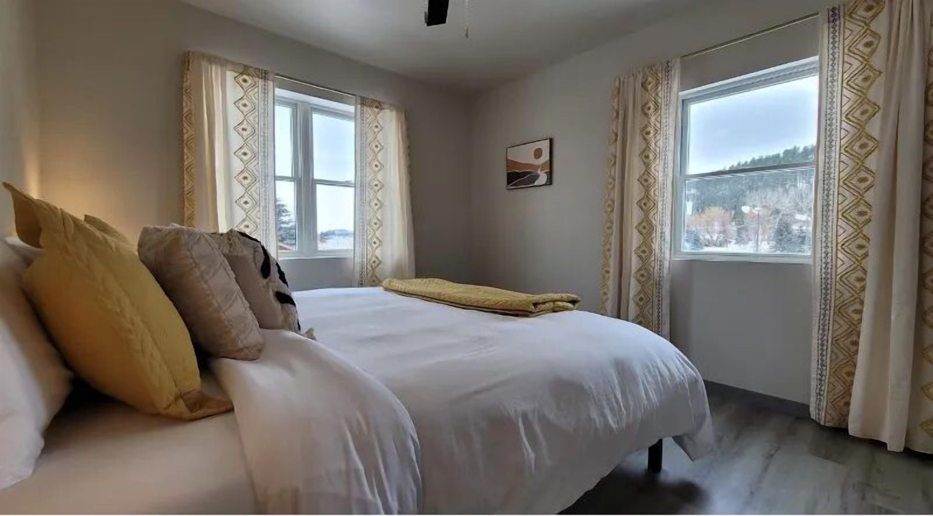 A bedroom with a large bed, two windows with patterned curtains, and a painting on the wall.