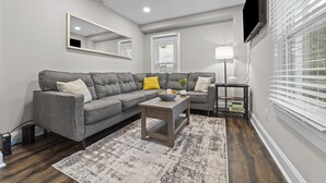 A modern living room with a grey sectional sofa, a small wooden coffee table, a floor lamp, and a flat-screen TV mounted on the wall. - Big House Downtown Greensboro (Greensboro)