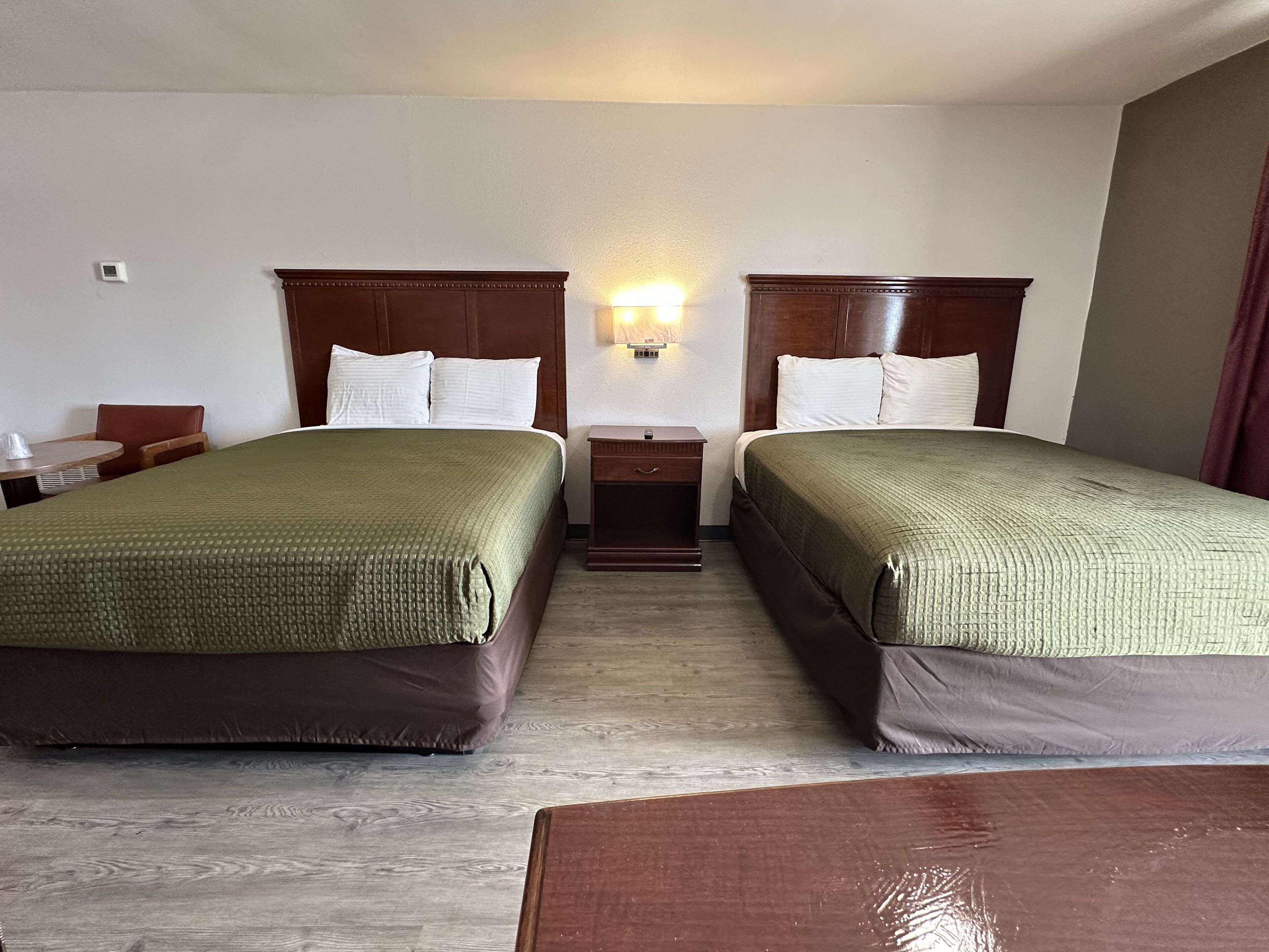 A hotel room with two beds, a nightstand, a lamp, and a small table.