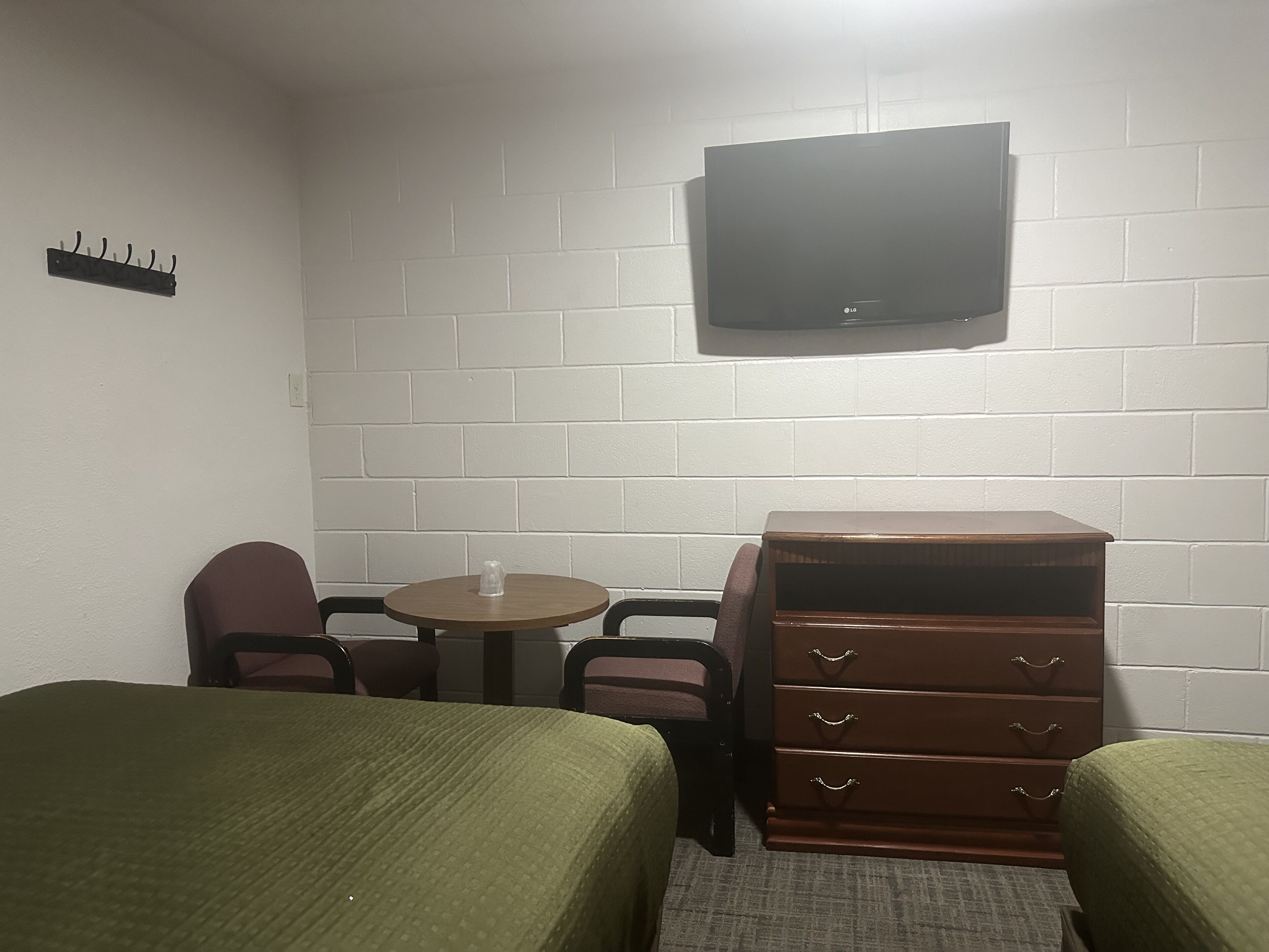 A small, single-room hotel room with two single beds, a wooden dresser, a small table, and a TV mounted on the wall.