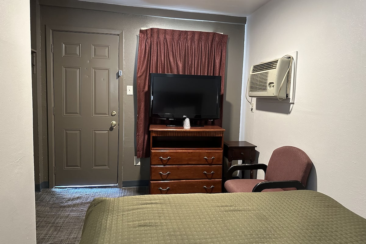 A hotel room with a bed, a wooden dresser, a television, a chair, and an air conditioning unit.