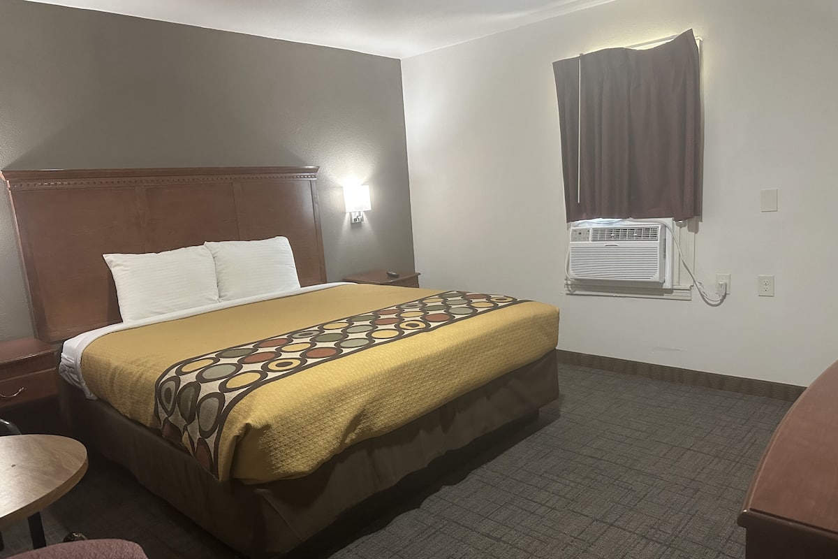 A hotel room with a bed, a desk, a chair, a window with curtains, and an air conditioning unit.