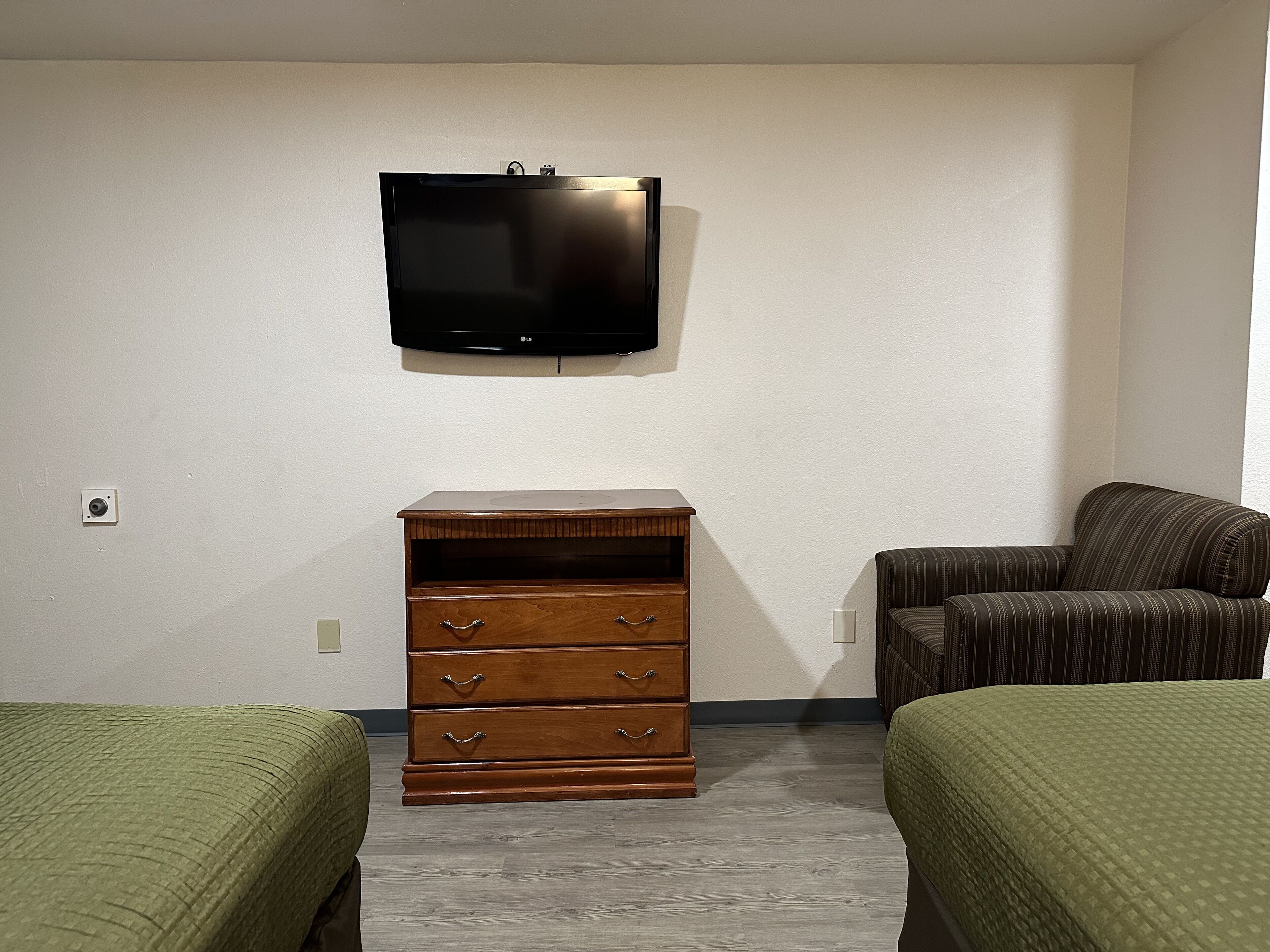 Una habitación con una televisión de pantalla plana montada en la pared, un aparador de madera con cajones, dos camas verdes y un sillón a rayas.