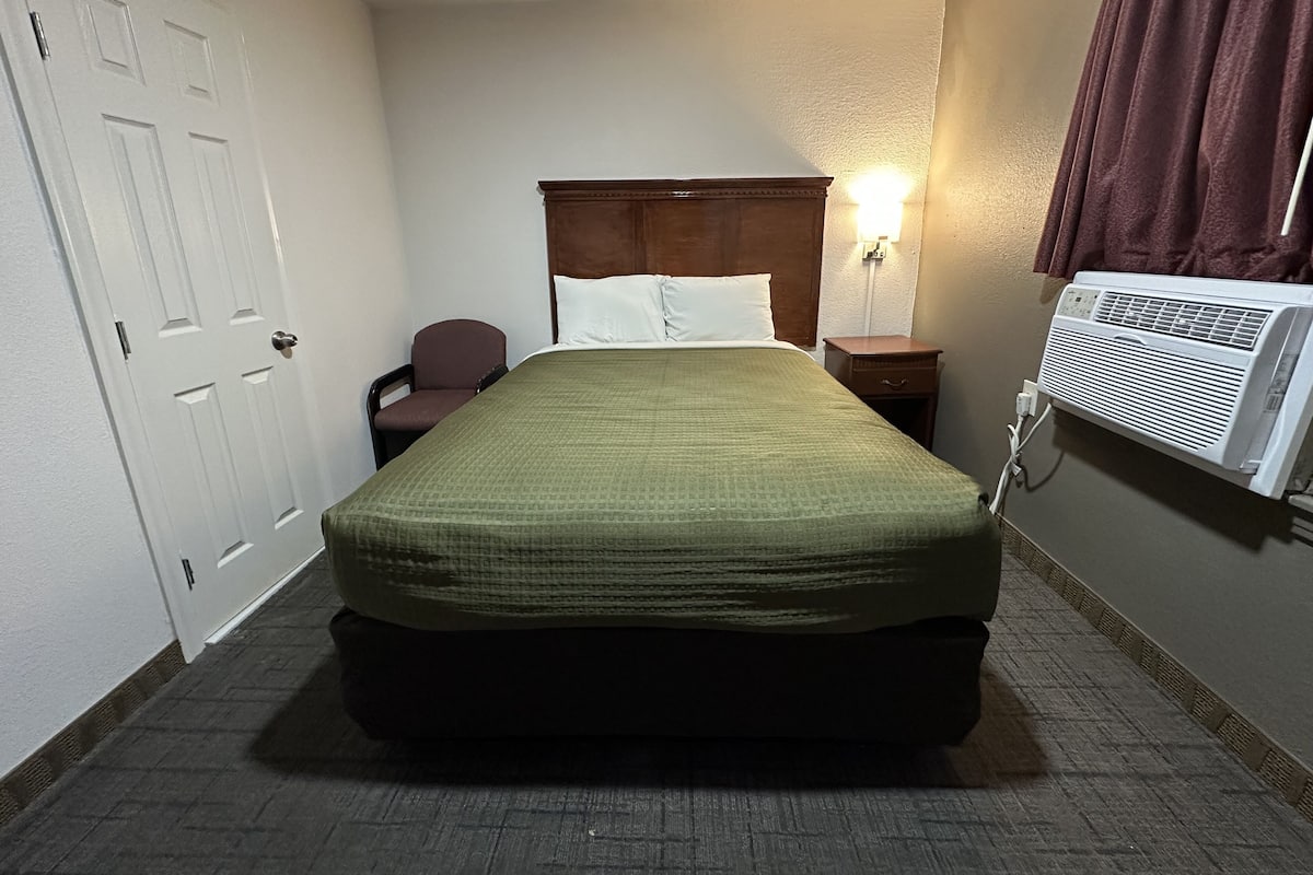 A hotel room with a bed, a chair, a nightstand, a wall-mounted air conditioner, and a door.