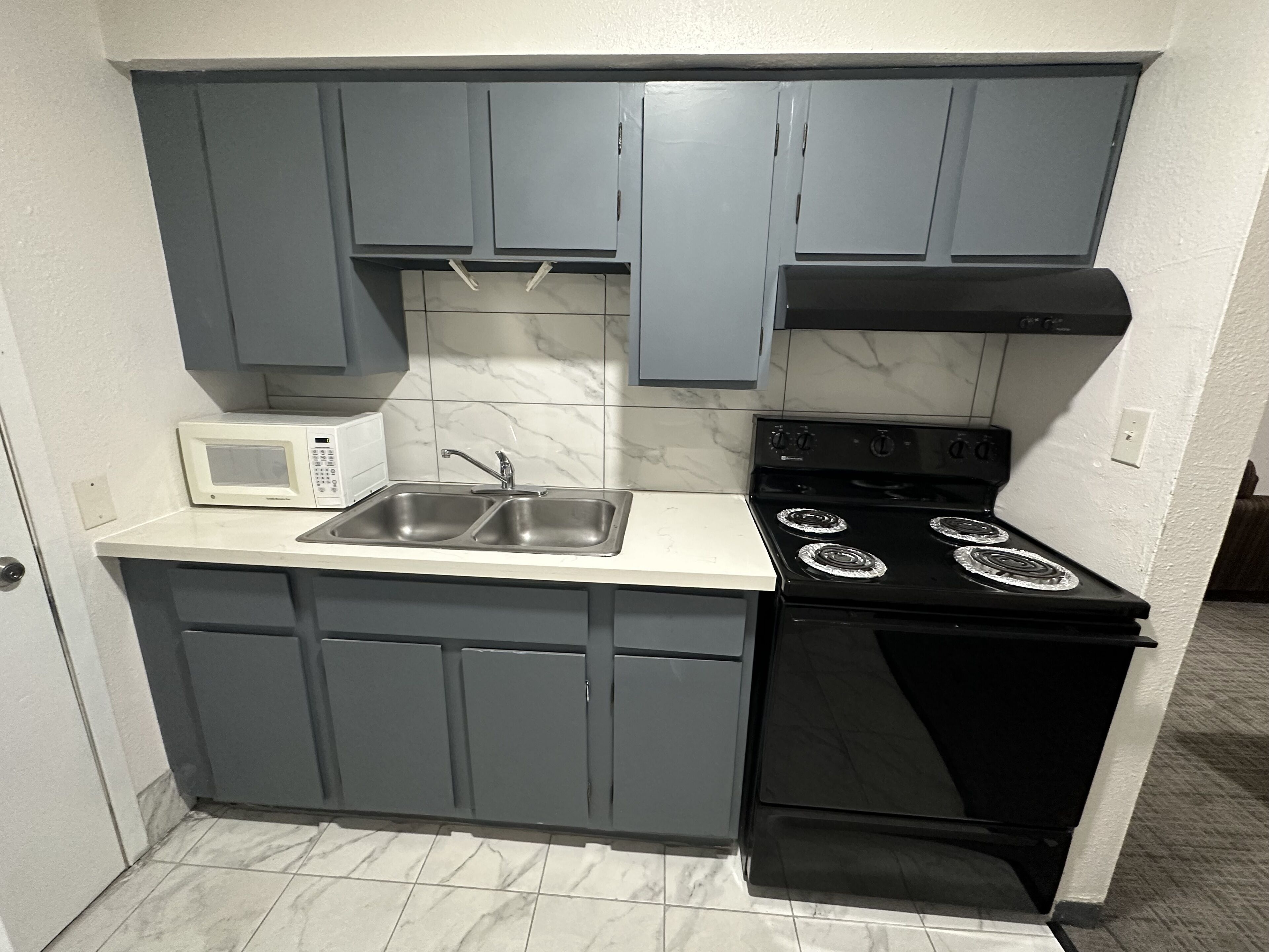 A compact kitchen with grey cabinets, a white countertop, a built-in microwave, a double sink, and a black stove with four burners.