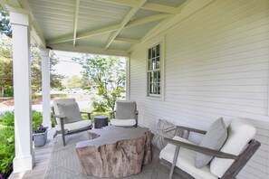A covered porch with a wooden table and four chairs, a window, and a view of a green landscape.