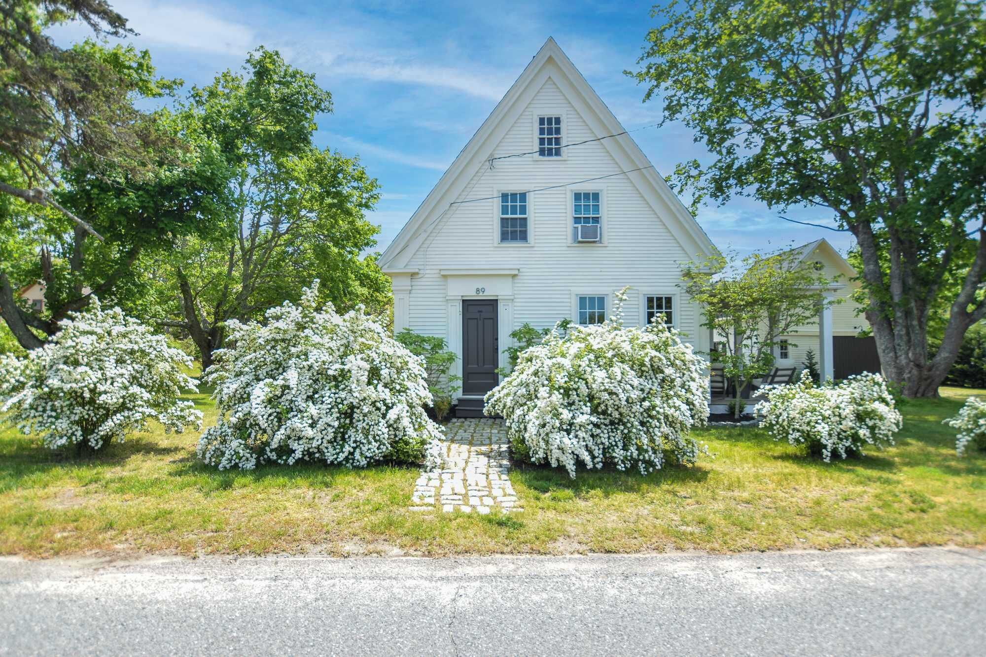 A white house with a triangular roof, a front door, and a number '89' on the door. There are three white flowering bushes in front.