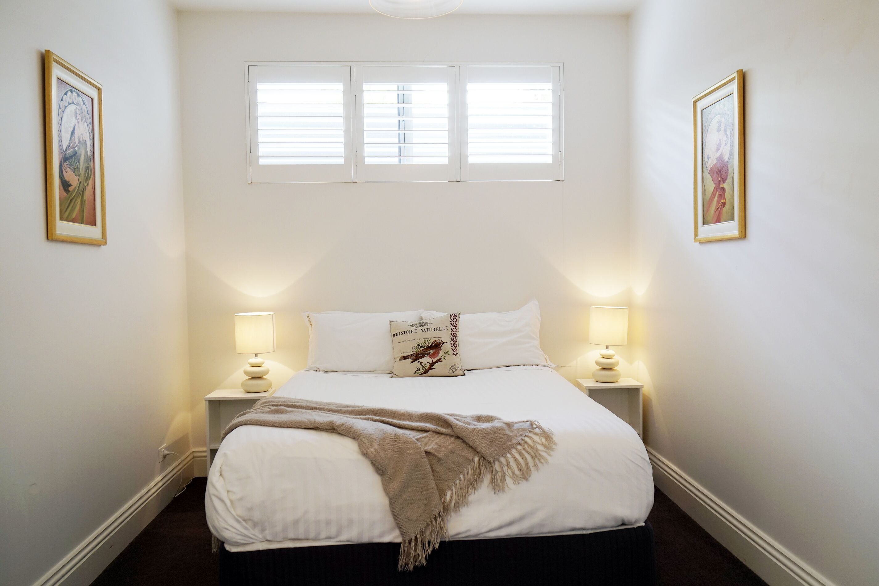 A neatly arranged bedroom with a bed, two bedside lamps, and two framed pictures on the walls.
