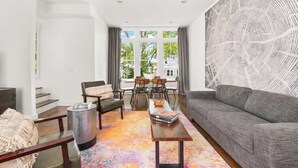 Un salon moderne avec un canapé gris, une table basse en bois et un tapis coloré. On y trouve également un coin repas avec une table et des chaises, ainsi qu’un escalier menant à un autre niveau.