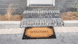 Una puerta blanca con un panel de vidrio vertical, un felpudo con la palabra “WELCOME” y una pared de ladrillo.