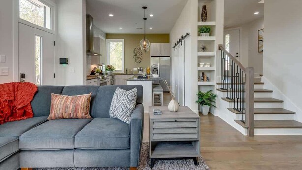 Un salón moderno con un sofá gris, una escalera y una cocina al fondo.