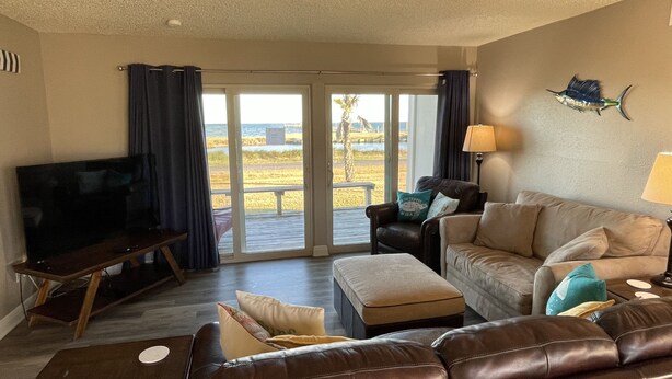 A living room with a television, leather sofas, a coffee table, and a view of the beach through large glass doors.