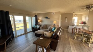A spacious living room with a large sofa, a coffee table, and a dining area with a table and chairs. There is a sliding glass door leading to a balcony with a view of the beach.