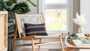 Ein Korbstuhl mit schwarzem Sitzkissen in einem Zimmer, das eine große Pflanze, ein Fenster und einen Teppich mit Webmuster beherbergt.