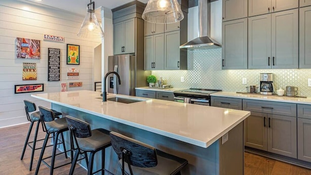 Een moderne keuken met grijze kastjes, een centraal eiland met barstoelen, hangende verlichting en een spatwand met een geometrisch patroon.