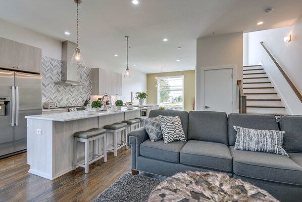 A modern kitchen with a central island, bar stools, and a grey sofa in a living area. - Group Getaway | Walkable Gulch Location + Parking! (Nashville)