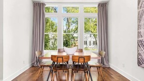 Un espace repas comprenant une table et des chaises, de grandes fenêtres avec des rideaux, et une vue sur la verdure et les bâtiments environnants.