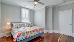 Une chambre à coucher comprenant un lit, un ventilateur de plafond, une fenêtre avec des stores, une table de chevet avec une lampe et une porte.