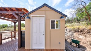 A small, single-story building with a white door and a window, located on a wooden deck with a pergola overhead.