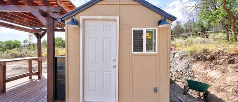 A small, single-story building with a white door and a window, located on a wooden deck with a pergola overhead.