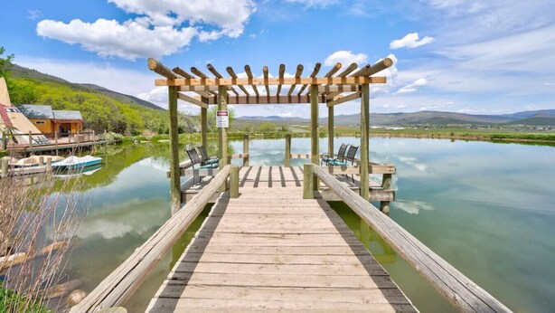 A wooden pier leading to a gazebo over a calm lake, with a boat docked nearby and a small building on the left.
